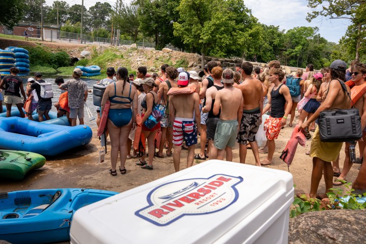 Group of people in swimwear gathered near river with rafts and trees.