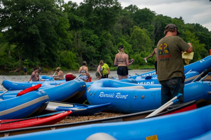 Group preparing inflatable blue rafts by a riverside with trees in the background.