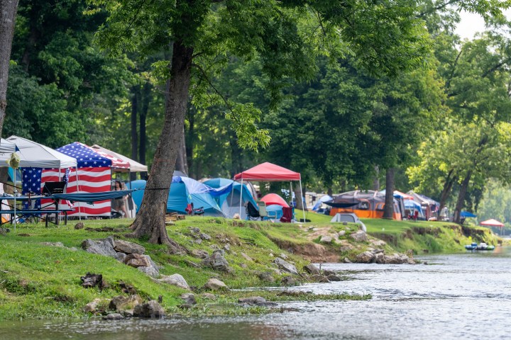 Campsite by a river with tents and trees under a clear sky.