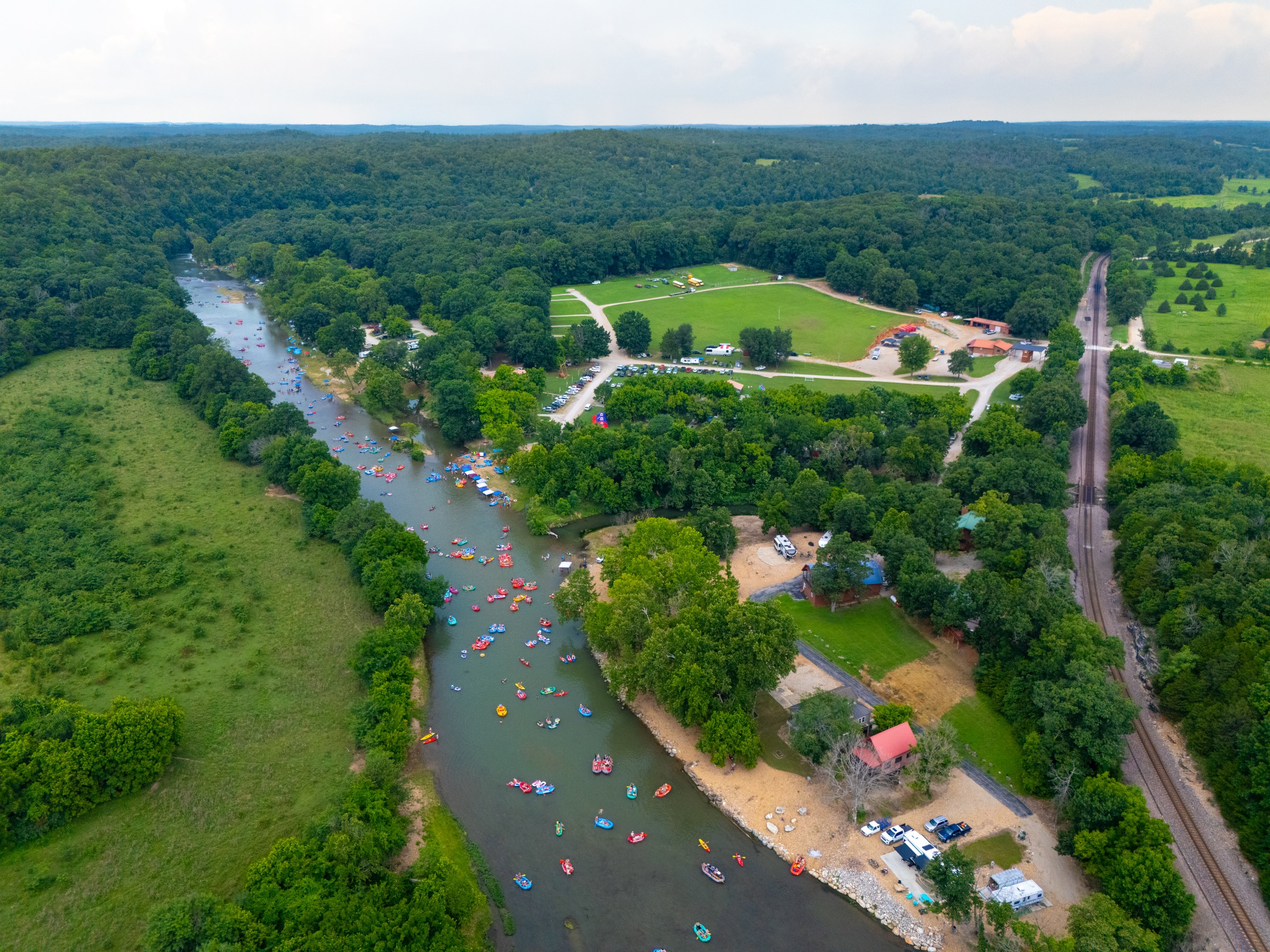 Aerial view of a river with many kayaks, surrounded by lush green trees and fields.