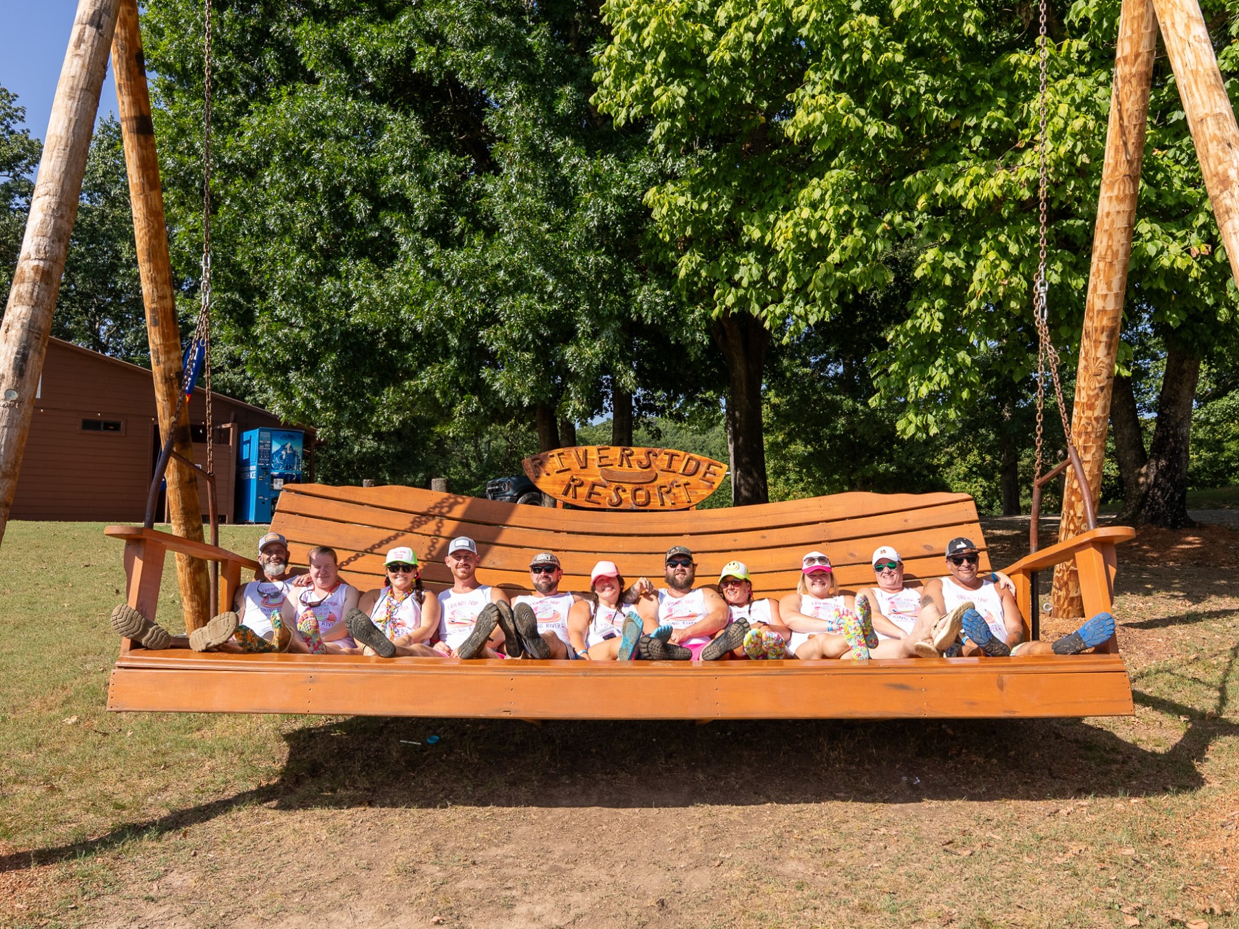 Group of people sitting on a large wooden swing labeled 'Riverside Resort' in a grassy area with trees.
