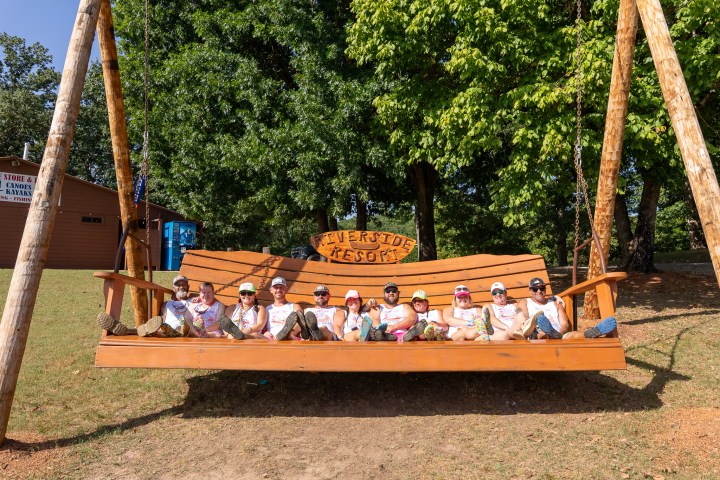 Group of people sitting on a large wooden swing labeled 'Riverside Resort' in a grassy area with trees.