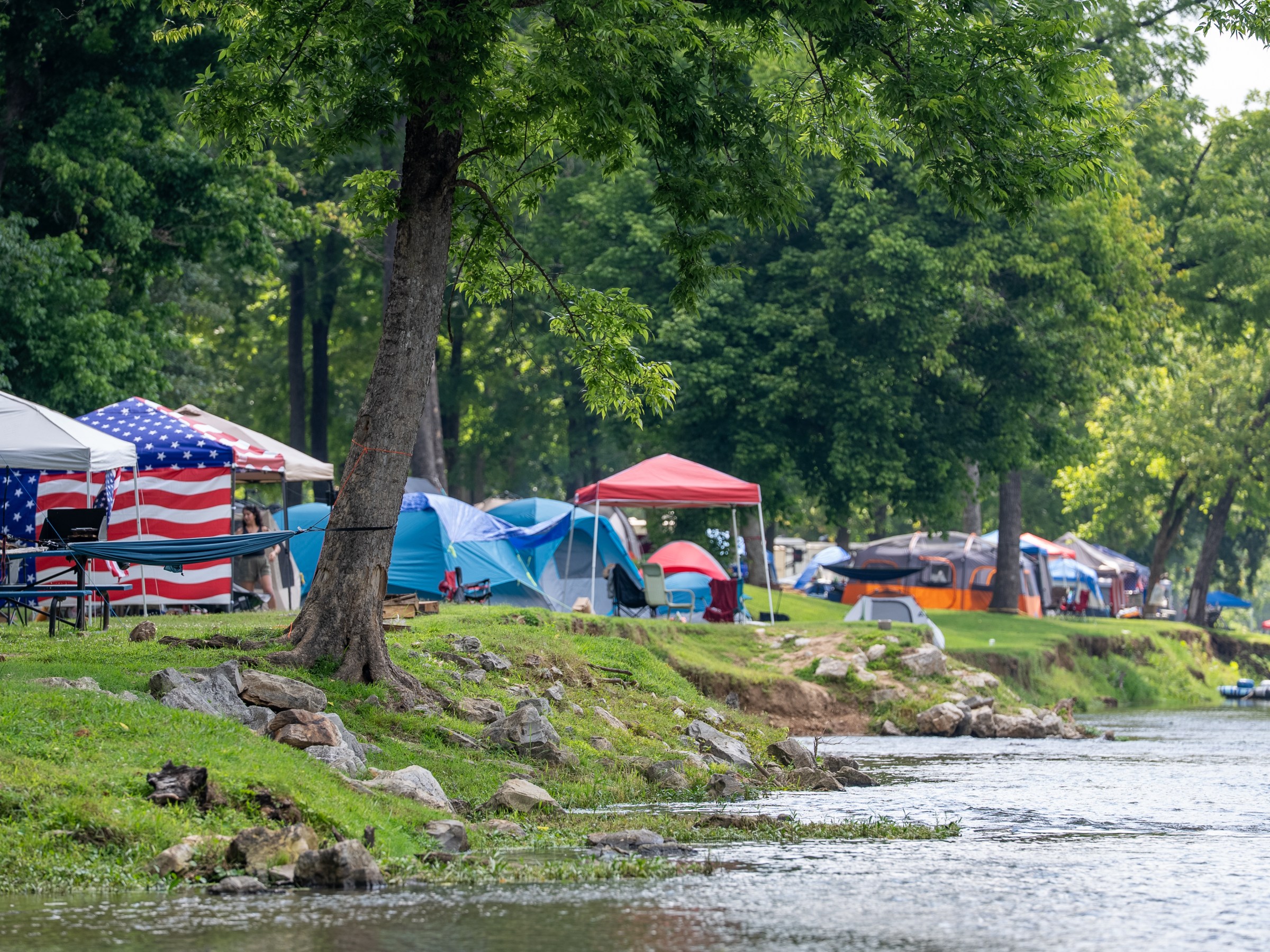 Campsite by a river with tents and American flag canopy.